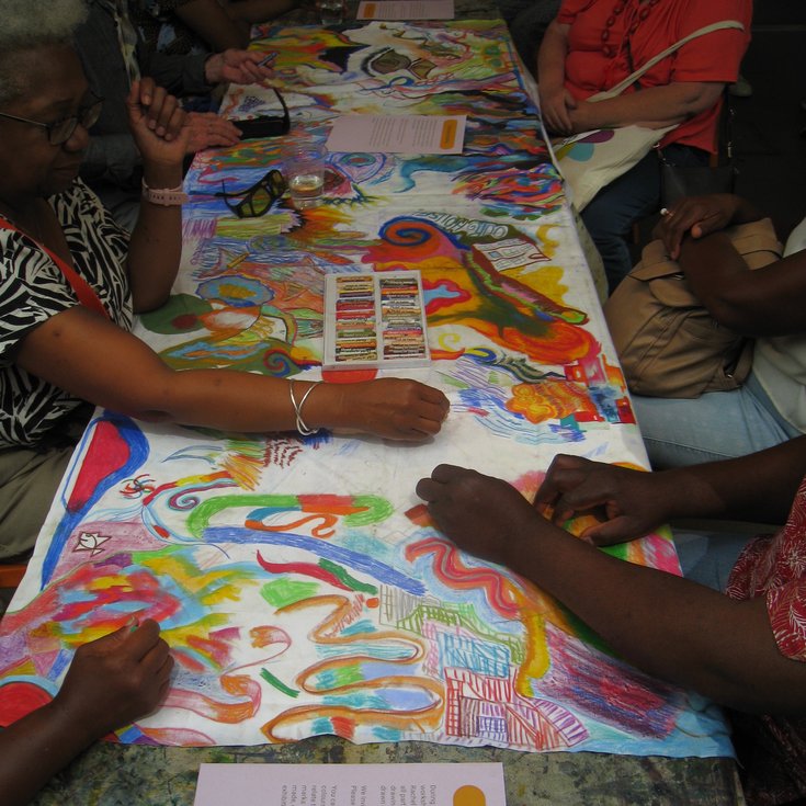 People taking part in an art workshop, lots of hands drawing colourful patterns on a giant white surface using oil pastels.