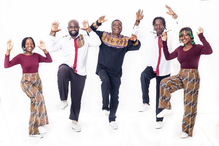 5 singers in a group with hands in the air and one leg lifted. Each singer wears an outfit of red, black and white