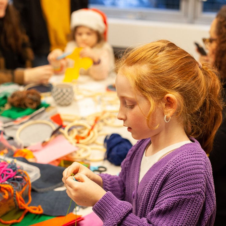 Young girl in a purple cardigan sitting at a table filled with colourful fabric scraps. She's holding a piece of thread.