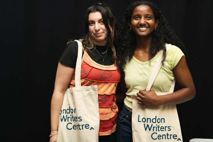 Two young women smile to camera carrying beige tote bags with the words London Writers Centre engraved on them.