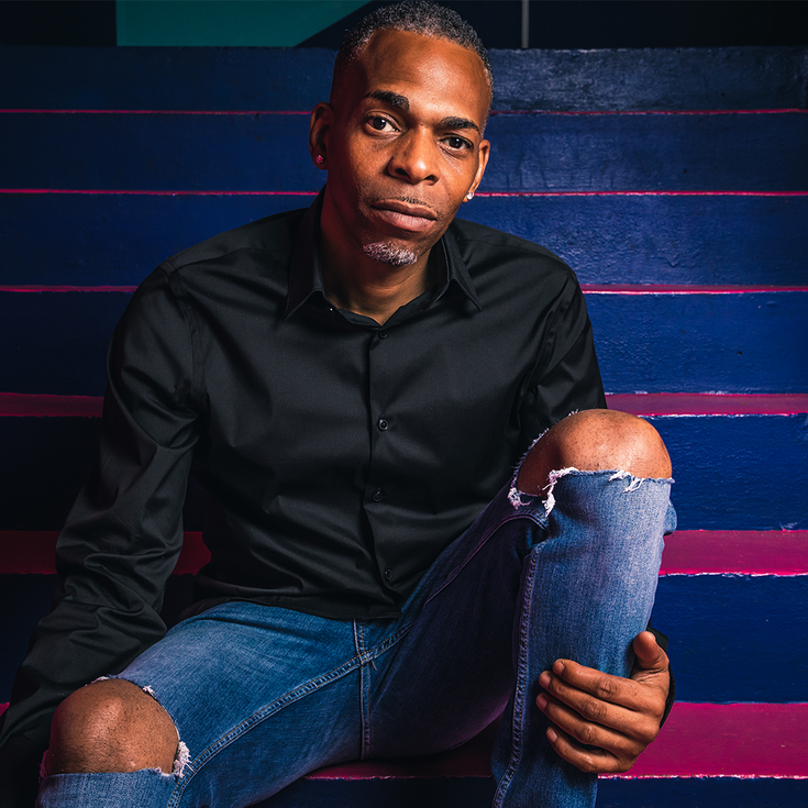 The comedian Slim sits on bright coloured stairs, he is wearing jeans with a rip in the knee and a black shirt. He sits on a step with his knee up and looks directly at the viewer.