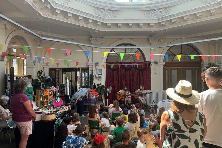 A crowd of people watching two people play the guitar and sing. They're inside a large open space with bright coloured bunting