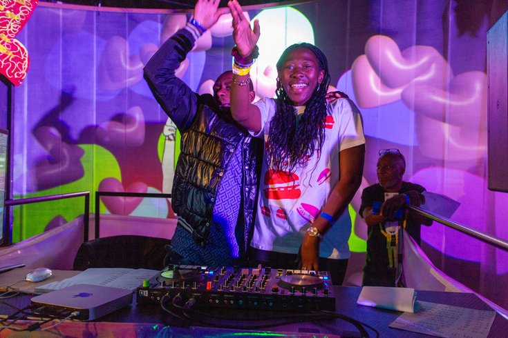 Black woman with mid length black plaits. She is wearing a white t-shirt with orange patterns. She is DJing and has her right hand in the air, she is smiling.