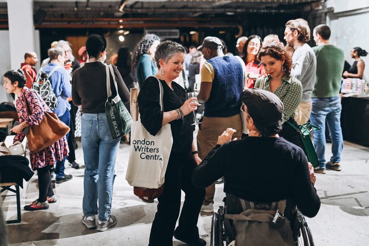 People chatting at an indoor networking event, including a wheelchair user speaking with two women, one holding a drink and a “London Writers Centre” tote bag.