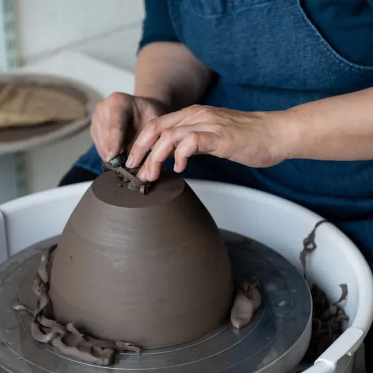 Maker's hands making a pot our of brown clay, gently touching the base of the pot turned upside down to refine it's base on a wheel.