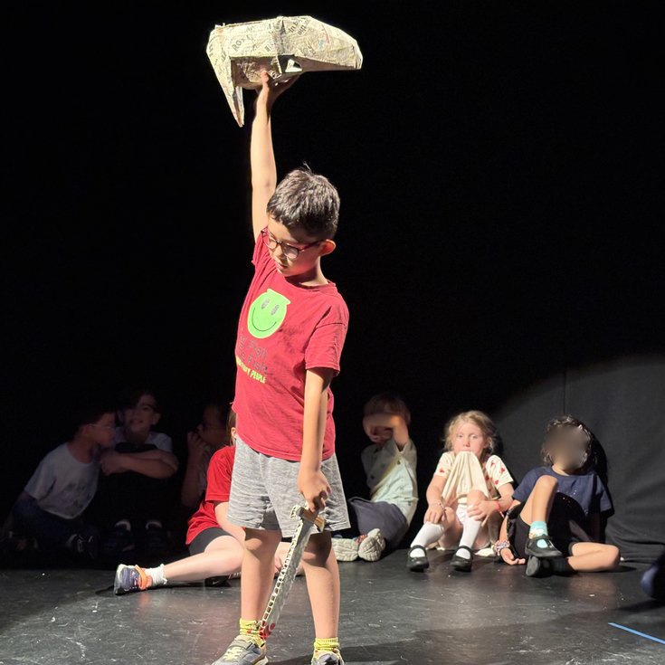 A student holding a sword and mask poses on stage as other students watch from the background.