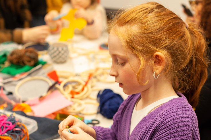 Young girl in a purple cardigan sitting at a table filled with colourful fabric scraps. She's holding a piece of thread.