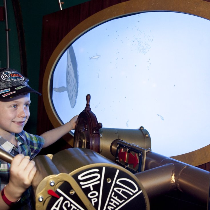 Young boy turning a submarine's controls, with a porthole window behind him showing an underwater scene