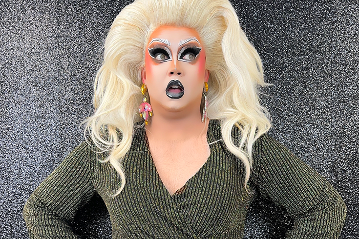 THATGIRL stands in front of a sparkly grey background, she looks off to the side with her mouth slightly open, she has big blonde hair, pink earrings, sparkly makeup.