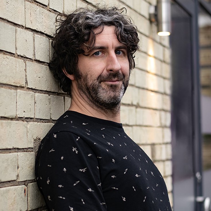 Male with dark hair and stubble wearing a black t-shirt leaning against a brick wall