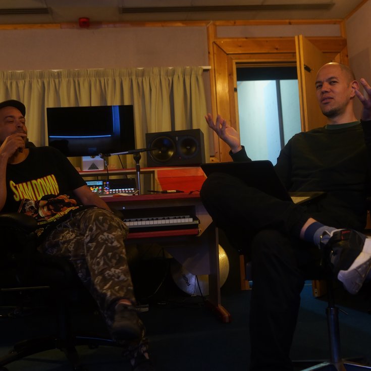 Two men sit in a music studio; the one on the left wears a cap, black top, and camo trousers, while the one on the right is dressed in a black jumper and trousers.