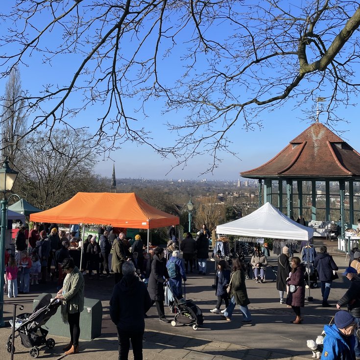 People visiting market stalls with the Horniman Bandstand in the background, with a blue sky and wintery trees above