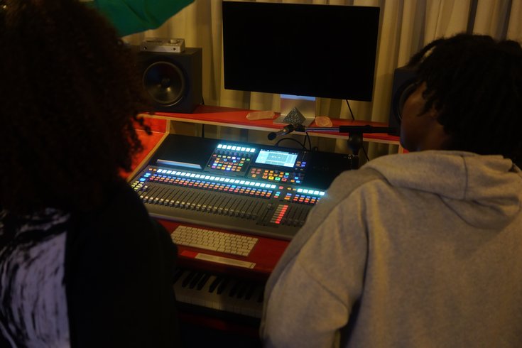 Two people, one in a grey jumper and one in a black jumper, are seen from behind as they look at a bright orange mixing desk in a music studio.