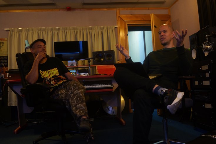 Two men sit in a music studio; the one on the left wears a cap, black top, and camo trousers, while the one on the right is dressed in a black jumper and trousers.