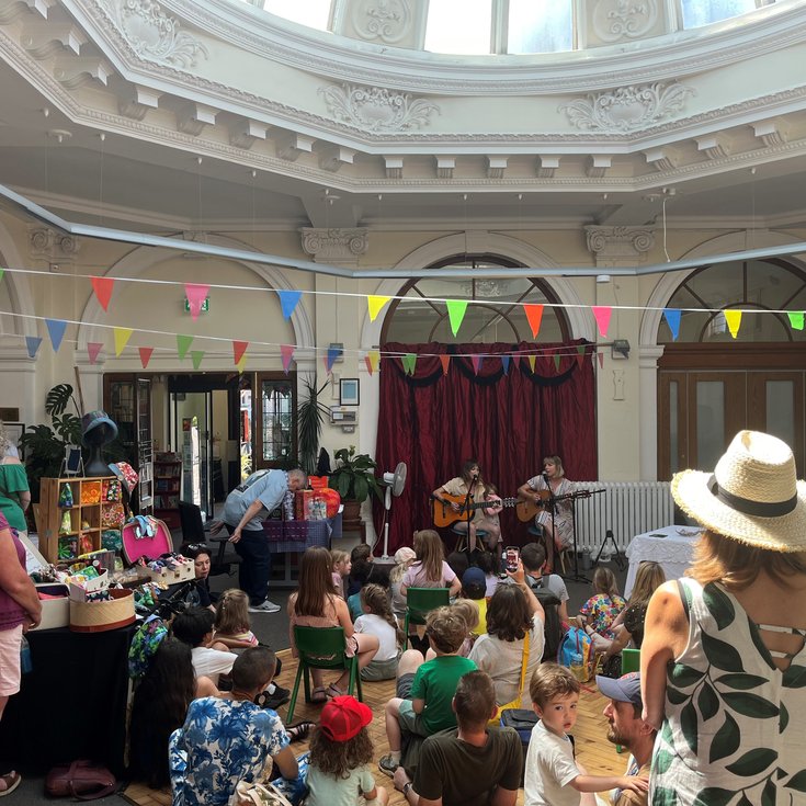 A crowd of people watching two people play the guitar and sing. They're inside a large open space with bright coloured bunting