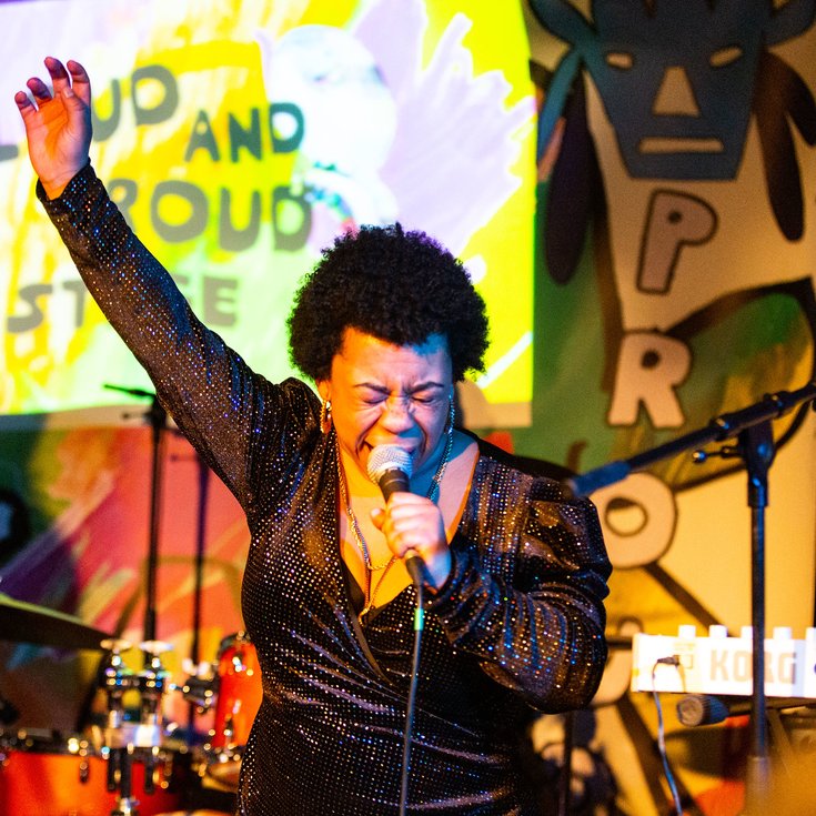 Black woman with short black hair wearing a black sparkly dress. She is singing into a mic with her right hand in the air and eyes closed.
