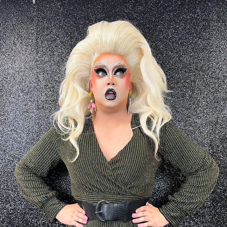 THATGIRL stands in front of a sparkly grey background, she looks off to the side with her mouth slightly open, she has big blonde hair, pink earrings, sparkly makeup.