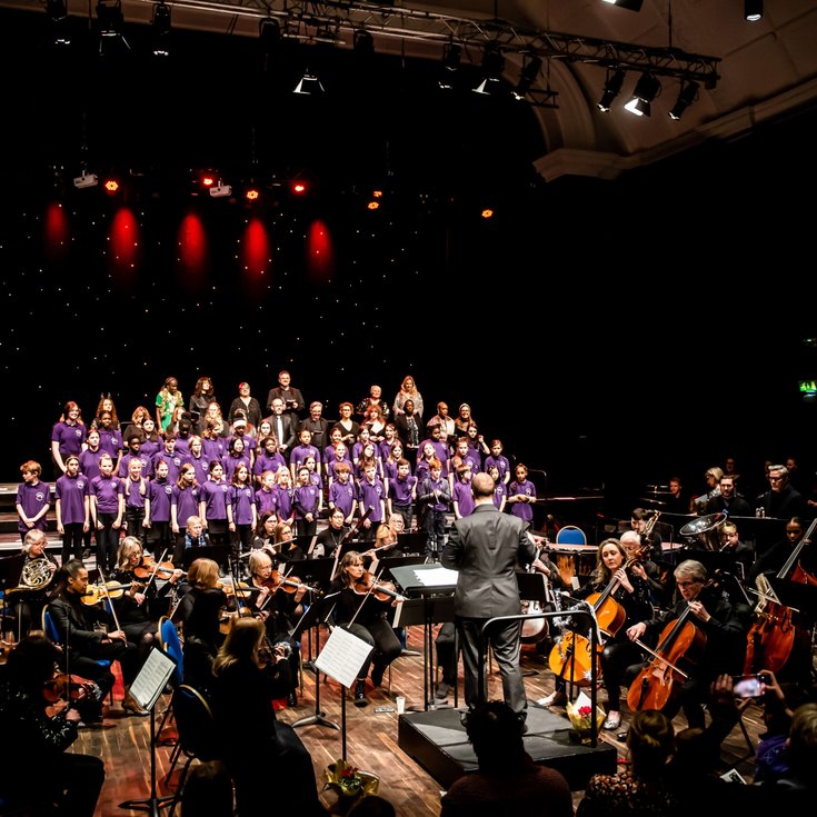 A number of people playing instruments as part of an orchestra and a choir singing on stage