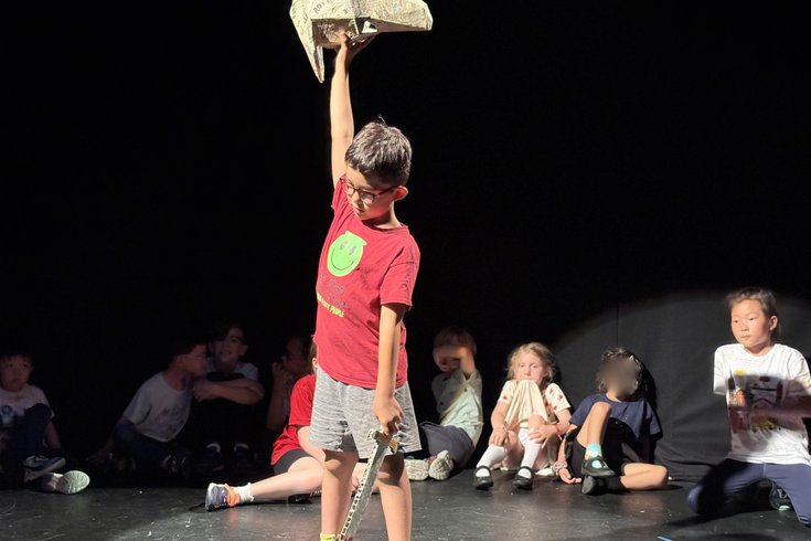 A student holding a sword and mask poses on stage as other students watch from the background.