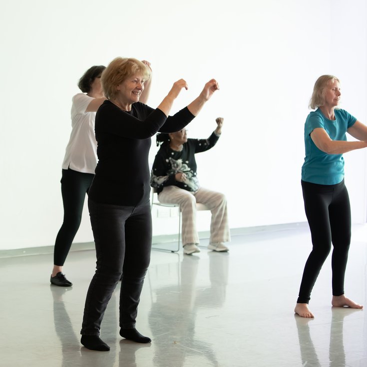 Group of over 60s participants  dancing together in a studio. One person is seated, some are standing.