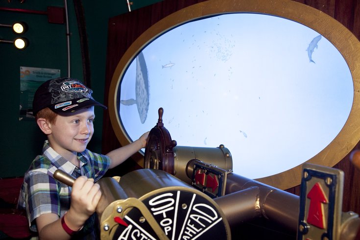Young boy turning a submarine's controls, with a porthole window behind him showing an underwater scene
