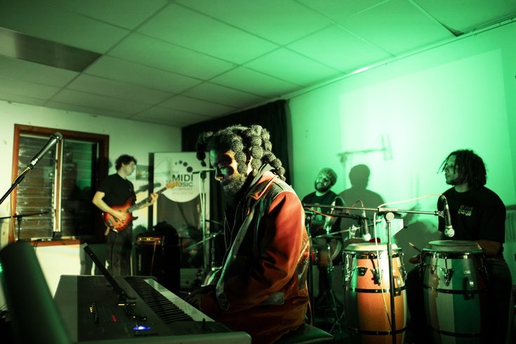 Headline performance by new patrons Yussef Dayes and Ahnansé from Steam Down with band members on piano, guitar, and drums under green lighting at The Midi Music Company’s 30th anniversary.