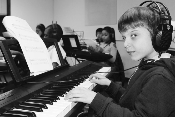 A black and white image of a young boy with a short haircut, wearing headphones, looks directly at the camera while playing a keyboard with a music sheet in a classroom full of children.