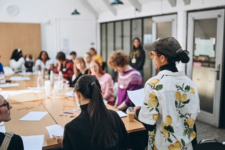 A group of people engaged in a workshop around a table, with one person standing and holding papers, in a bright, casual setting.