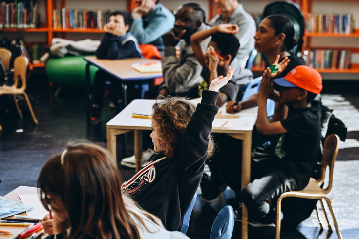 Children in a classroom, some raising hands, with books on tables.