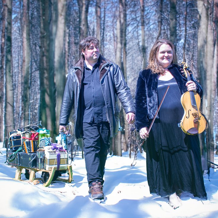A scene of a forest with snow on the ground. A man pulls a sleigh and a woman holds a fiddle
