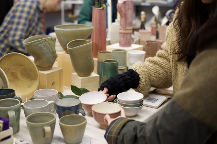 Visitors standing in front of a a display of handmade pottery. Vessels in green, pink and orchre glaze are carefully arranged on the workbench in front of them.