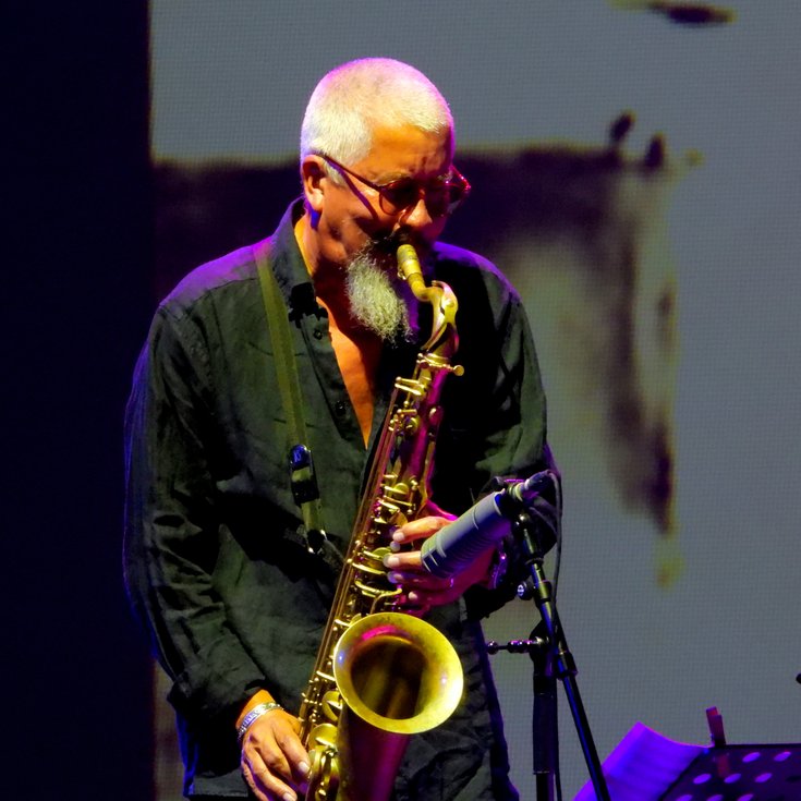 Male with white hair and a beard wears a black shirt and is playing the saxophone