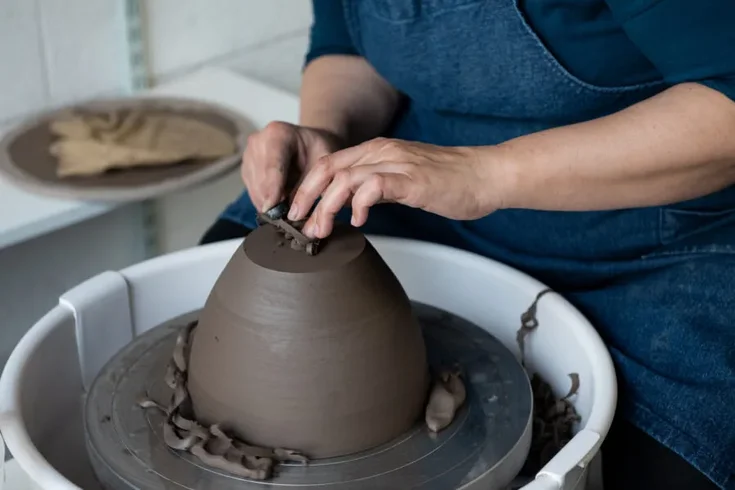 Maker's hands making a pot our of brown clay, gently touching the base of the pot turned upside down to refine it's base on a wheel.