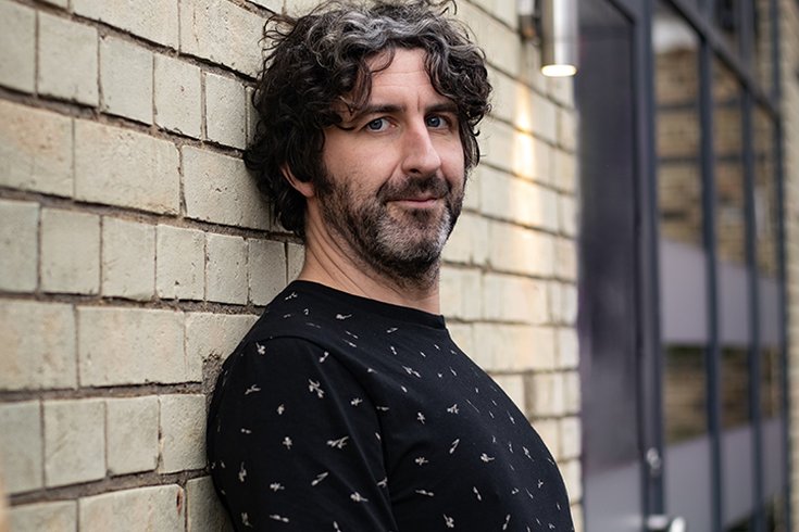 Male with dark hair and stubble wearing a black t-shirt leaning against a brick wall