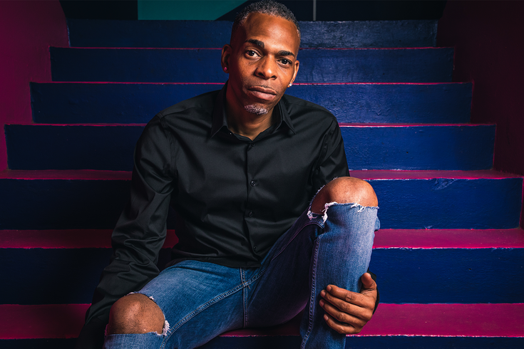 The comedian Slim sits on bright coloured stairs, he is wearing jeans with a rip in the knee and a black shirt. He sits on a step with his knee up and looks directly at the viewer.