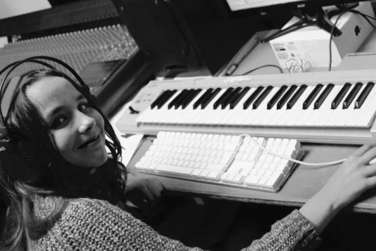 A young girl with straight, medium-length hair sits at a desk, smiling as she wears headphones and clicks a computer mouse beside a keyboard. She is dressed in a cozy knitted jumper.