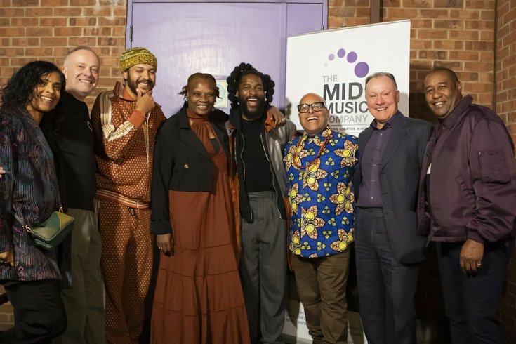 The founder, board members, and patrons of The Midi Music Company smile and laugh while posing for a photo at the 30th anniversary celebration. A banner behind them reads "The Midi Music Company."