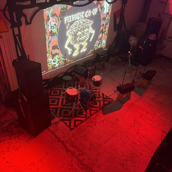 A railway arch, with a projection screen, PA system, and monitors set up. Lit by red light, with a projection saying 'Piehouse Co-Op' and a drawing of a pie man.