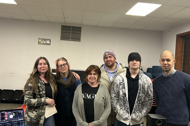 A group of six people stand behind DJ decks in a room, smiling and posing for a photo.