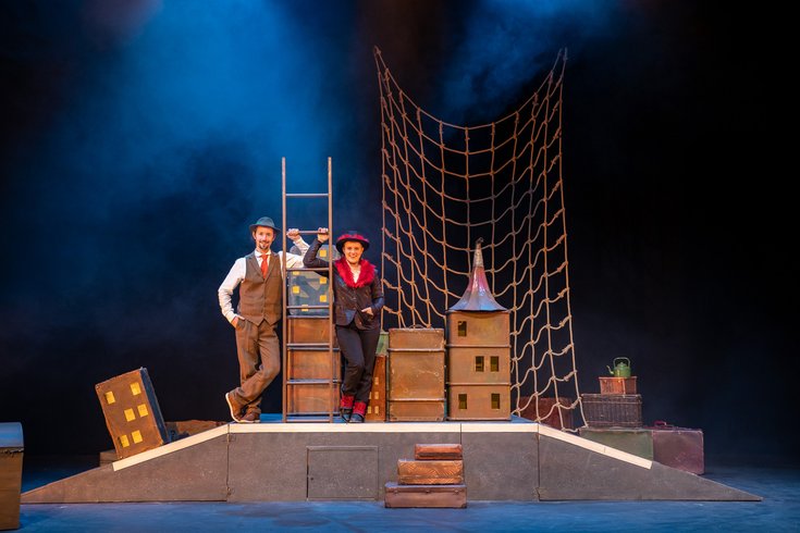 Two performers pose leaning on a ladder, with a net and boxes around them