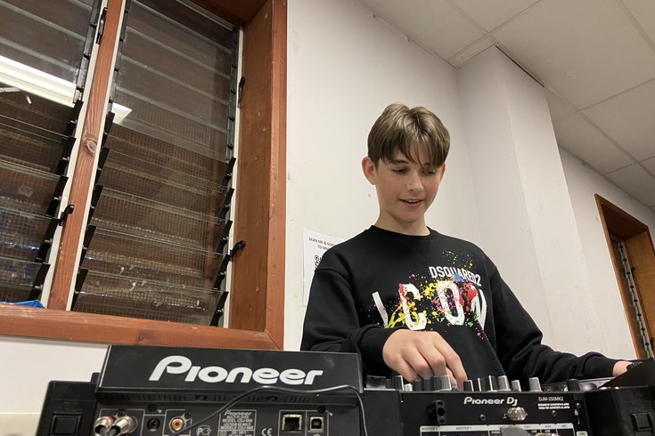 A young man with short brown hair, wearing a black jumper with a colourful paint-splatter design, mixes on black Pioneer-branded DJ decks.