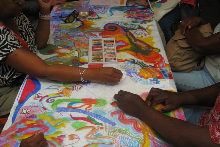 People taking part in an art workshop, lots of hands drawing colourful patterns on a giant white surface using oil pastels.