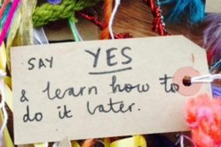 A brown luggage tag is surrounded by brightly coloured craft materials and on it is written the words ' Say YES & learn how to do it later.' Credit: Fun Palaces