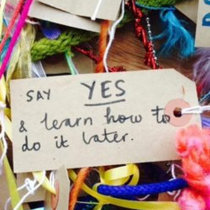 A brown luggage tag is surrounded by brightly coloured craft materials and on it is written the words ' Say YES & learn how to do it later.' Credit: Fun Palaces