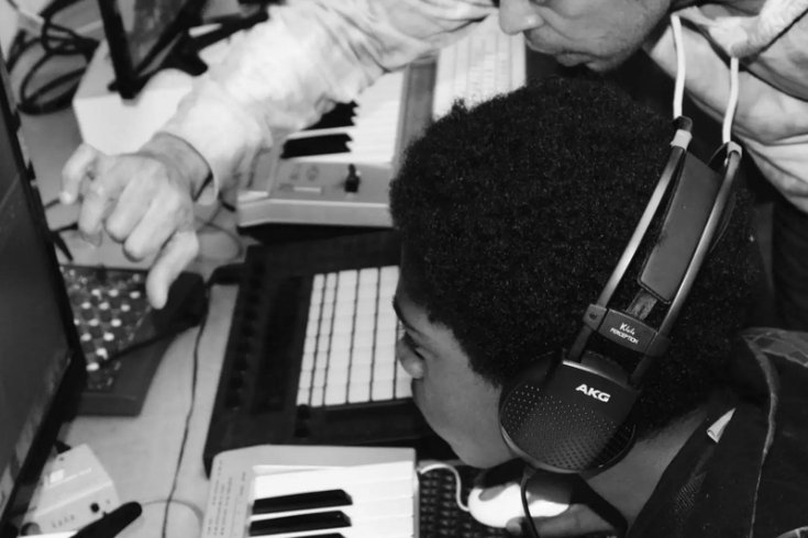 A black and white image of a boy wearing headphones sits at a keyboard and computer as a man points at the screen, guiding him.