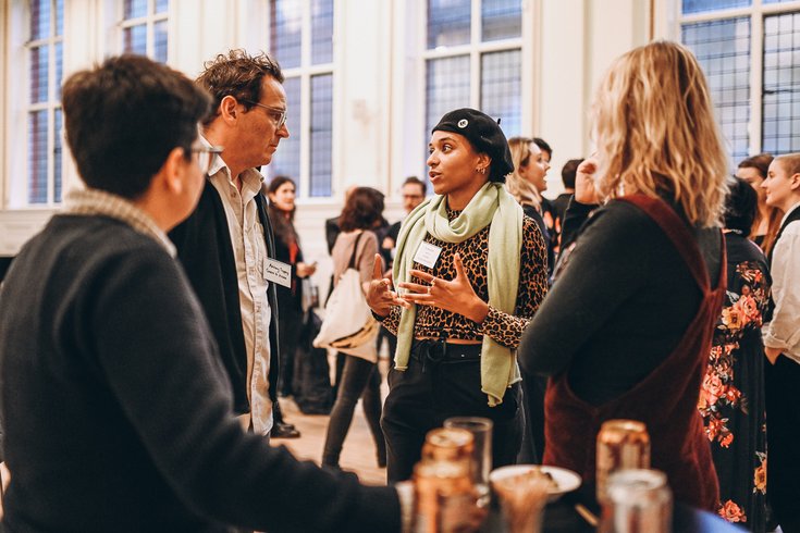 A group of people engaged in conversation in a well-lit room with tall windows.