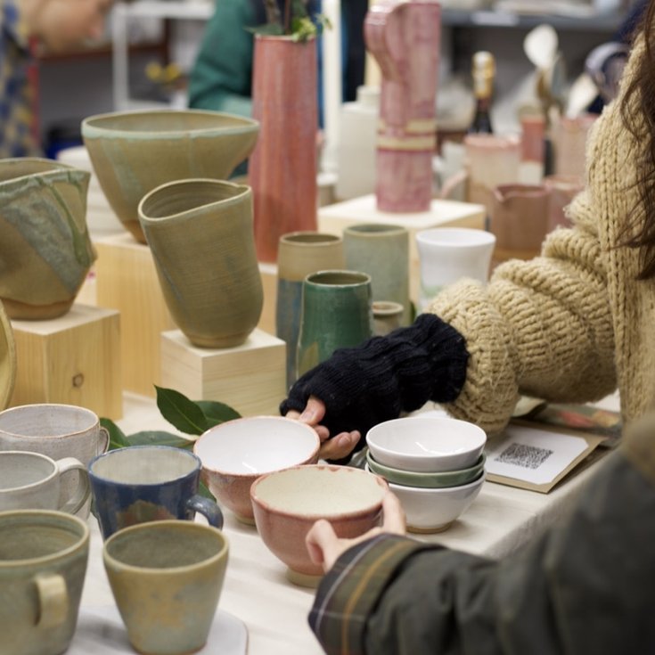Visitors standing in front of a a display of handmade pottery. Vessels in green, pink and orchre glaze are carefully arranged on the workbench in front of them.