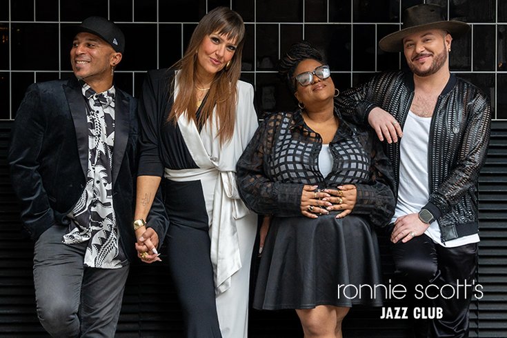 two females and two male singers all wearing black and white clothes standing against a black tiled wall