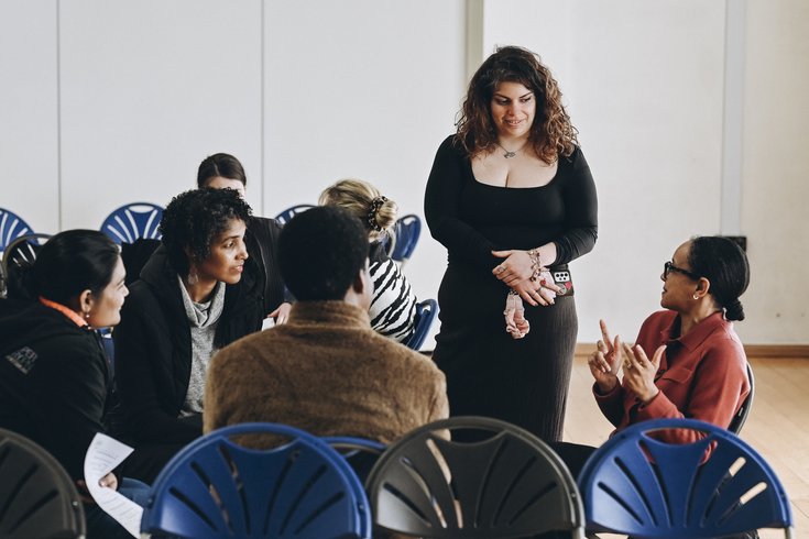 Small group discussion during a workshop or community event, with participants seated in a circle and engaged in conversation. One woman stands listening while another gestures as she speaks.