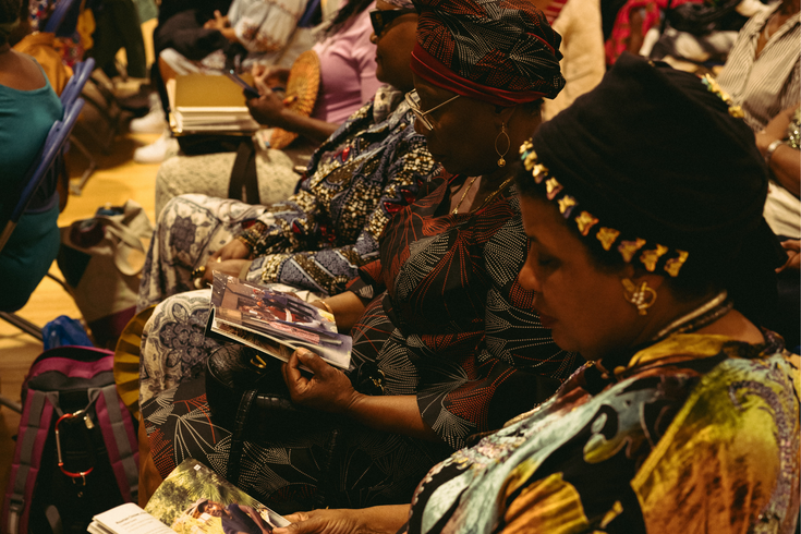 Several women in patterned attire sit in rows, attentively reading booklets or magazines during an indoor event or gathering.